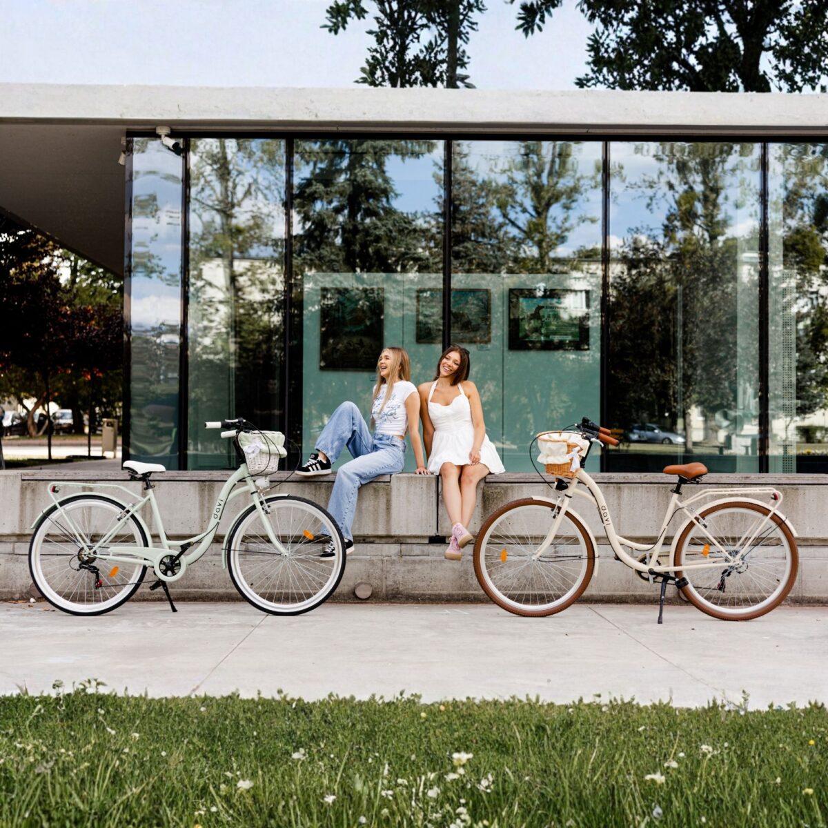 Kaksi naista istuu, polkupyörät edessä lasirakennus.